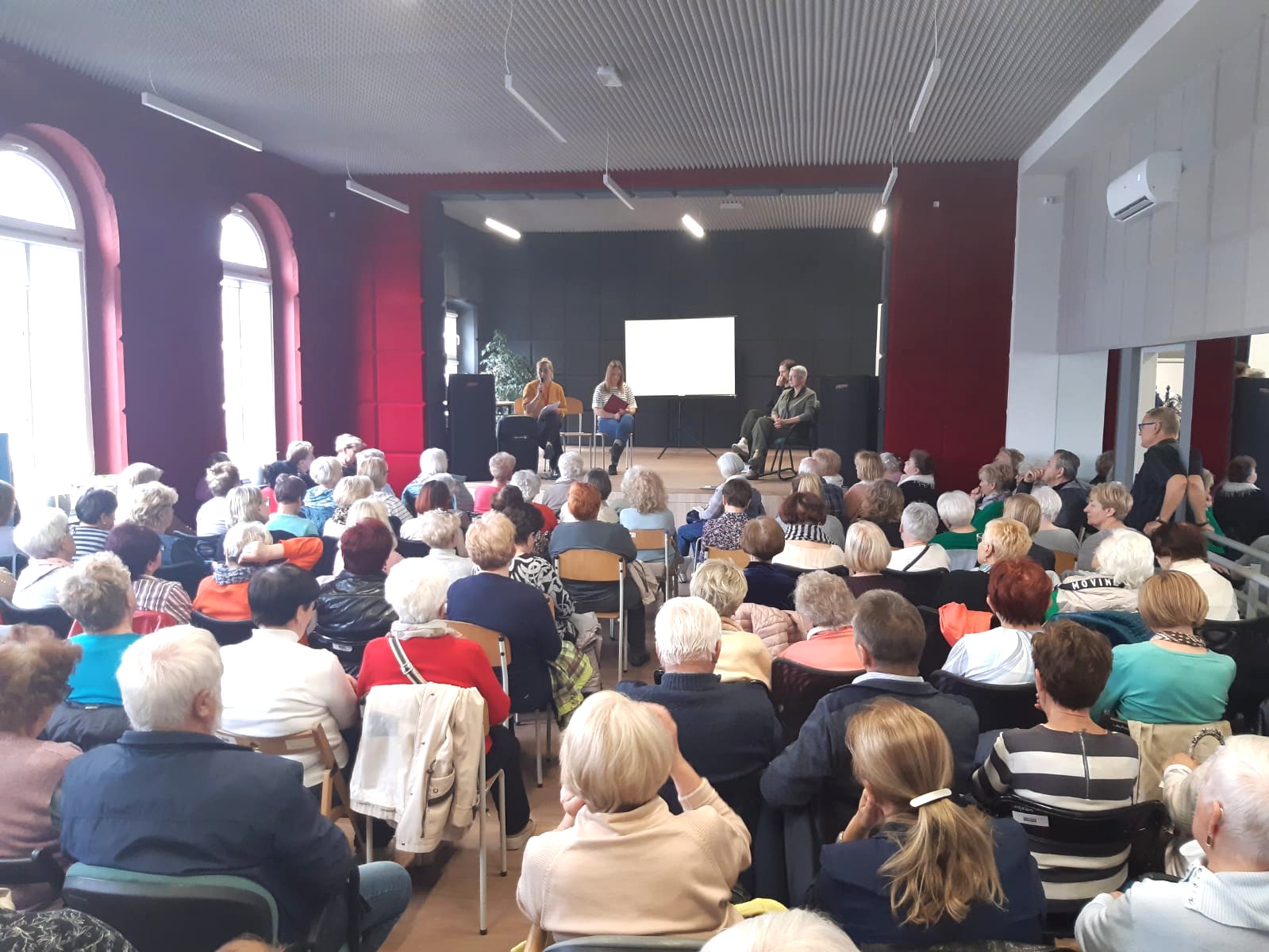 Zdjęcie przedstawia spotkanie organizacyjne dla seniorów w sprawie nowego programu usług społecznych i swarzędzkiej karty mieszkańca. Widać salę wypełnioną siedzącymi seniorami, a na scenie siedzą prelegentki przekazujące informacje zainteresowanym.