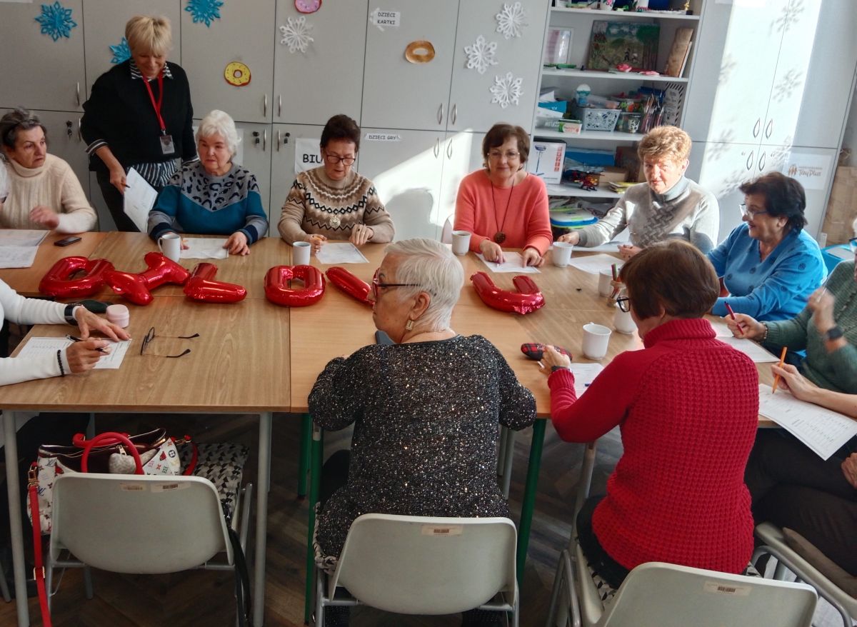 Grupa seniorek uczestniczy w zajęciach przy stole ozdobionym balonami z napisem 'LOVE'