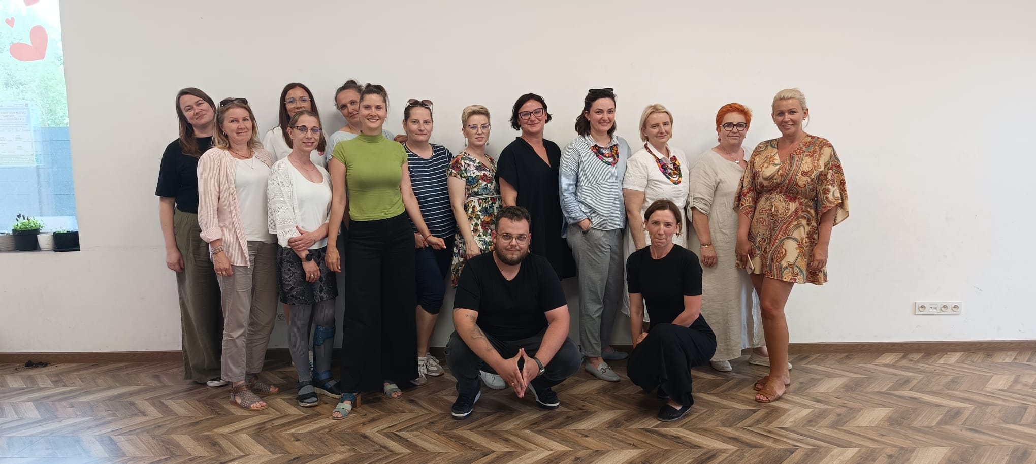 Zdjęcie wykonane w Centrum Międzypokoleniowym Pogodna. W sali na tle ściany stoi trzynaście przedstawicielek kół gospodyń wiejskich, kuca jeden mężczyzna również członek jednego z kół oraz organizatorka społeczności lokalnej.