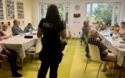 Zdjęcie przedstawia grupę seniorów siedzących przy stolikach zwróconych do policjantki, która stoi na środku pomieszczenia.