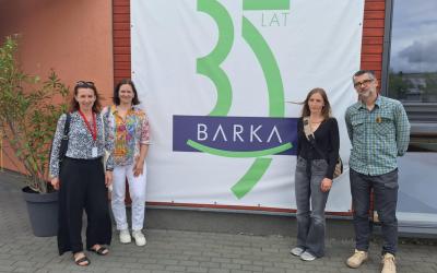 Czworo uczestników spotkania stoi na tle banera z napisem Barka 35 lat. Baner wisi na ścianie budynku, który jest siedzibą Fundacji Barka w Poznaniu.