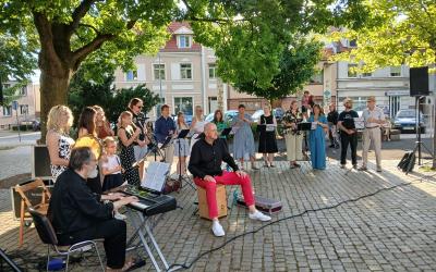 Plac Niezłomnych. Swarzędzka Orkiestra Flażoletowa daje koncert plenerowy na Placu Niezłomnych pod dużym dębem. W tle widać drzewa, kamienice i samochody.