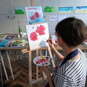 Zdjęcie przedstawia kobietę z paletą malarską malującą pędzlem na płótnie czerwone maki. Płótno stoi na sztaludze. Za nim znajduje się wzór obrazu.