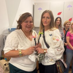 Dwie uśmiechnięte kobiety trzymają w dłoniach kwiaty zrobione na szydełku. W tle więcej kobiet.