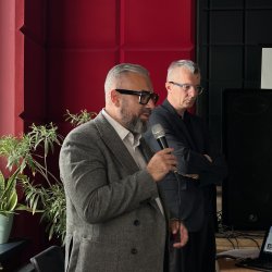 Dwóch mężczyzn stoi przed sceną w sali Centrum Aktywności Seniora. Jeden z nich mówi do mikrofonu.