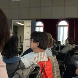 Zdjęcie przedstawia widok boczny osób siedzących na widowni. W tle widać lustra, w których odbijają się okna z przeciwległej ściany.