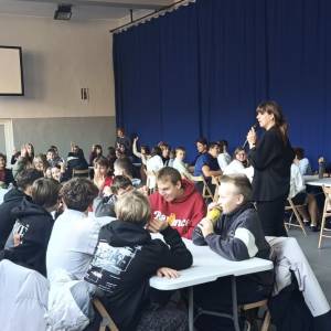 Zdjęcie przedstawia spotkanie z uczniami w sprawie utworzenia Rady Młodzieży. Jedna z prelegentek stoi pomiędzy uczestnikami i mówi do mikrofonu.