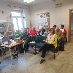 Zdjęcie wykonane od strony prowadzącego. Widać na nim siedzących i słuchających uczestników. Tło stanowi ściana z tablicą korkową i okno pełne światła.