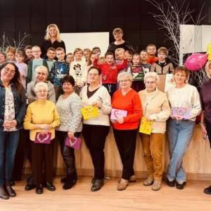 Zdjęcie grupowe wykonane w Centrum Aktywności Seniora. Przed sceną stoją seniorzy i pracownicy centrum. Na scenie stoją dzieci i panie z przedszkola.