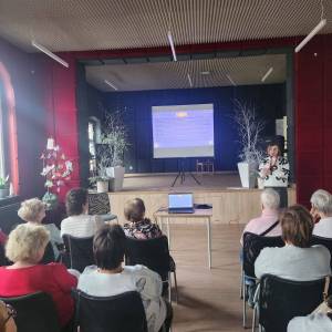 Główna sala w Centrum Aktywności Seniora. Na scenie stoi ekran, przed sceną stoi stół z projektorem oraz prelegentka, która mówi do audytorium.