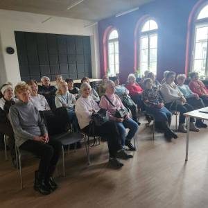 Główna sala w Centrum Aktywności Seniora. Publiczność składająca się z kilkudziesięciu siedzących seniorów słucha wykładu na temat karnawału.