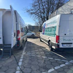 Zdjęcie przedstawia dwa samochody typu transporter służące do przeprowadzania badań serca i skóry stojące na parkingu przed Centrum Usług Społecznych.