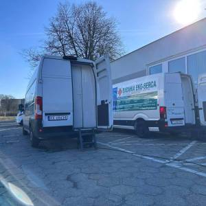 Zdjęcie przedstawia dwa samochody typu transporter służące do przeprowadzania badań serca i skóry stojące na parkingu przed Centrum Usług Społecznych.