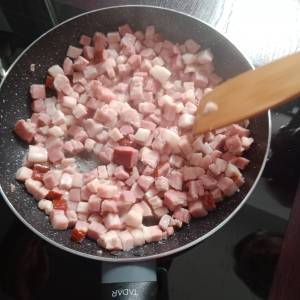 Zdjęcie przedstawia proces smażenia boczku pokrojonego w kosteczkę na dużej patelni. Widać też drewnianą szpatułkę dotykającą boczek.