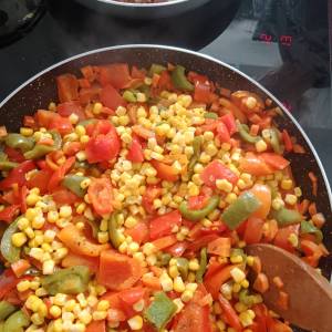 Zdjęcie przedstawia zbliżenie na patelnię, na której są różne pokrojone warzywa: czerwona i zielona papryka, pomidory i kukurydza.