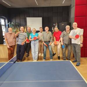 Grupa seniorów i jedna seniorka z rakietkami do tenisa stołowego pozują do zdjęcia w Centrum Aktywności Seniora. Zdjęcie wykonano zza stołu tenisowego.