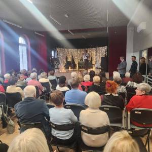 Seniorzy usadzeni w sali oglądają przedstawienie na scenie