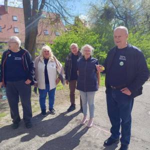 Podwórko przed Dziennym Domem Senior Wigor w słońcu. Grupa pięciorga seniorów wykonuje jedną z konkurencji marsz z jajkiem na łyżeczce na czas.