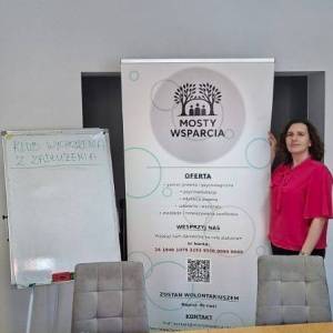  Zdjęcie ukazuje stół, na którym stoją tale z ciastkami i leżą inne przedmioty. Za stołem widać dwa krzesła. Za nimi przy rolapie stoi pani prawnik.