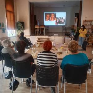 Sala główna Centrum Aktywności Seniora. Wolontariuszka z Francji wyświetla seniorom prezentację multimedialną o swojej rodzinie i miejscu zamieszkania.