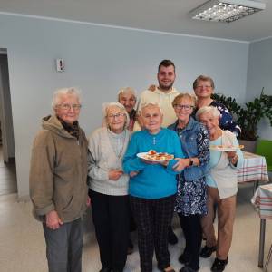 Wnętrze Dziennego Domu Senior Wigor. Uśmiechnięty wolontariusz stoi w otoczeniu uśmiechniętych seniorek. Dwie z nich trzymają talerze z kanapkami.