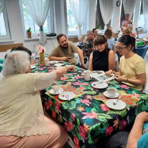 Dzienny Dom Senior Wigor. Wolontariuszka, pracownicy i seniorzy siedzą przy stole. Jedna z seniorek wskazuje coś w książce, którą trzyma wolontariuszka.