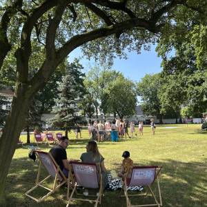Śniadanie na trawie. Widok na park. Widać piknikowiczów na leżakach, grupy dzieci bawiących się i wykonujących różne zadania.