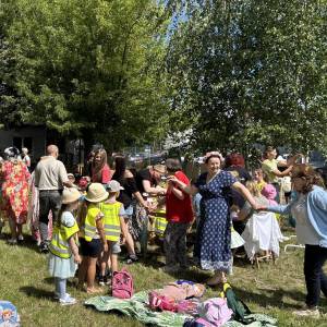 Powitanie lata w ogrodzie społecznym. Trwa zabawa. Pomiędzy kocami a stanowiskiem z warkoczykami seniorzy przechodzą łańcuszkiem, trzymając się za ręce.