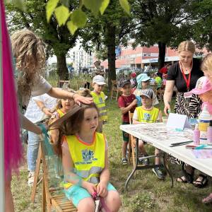 Powitanie lata z Centrum Usług Społecznych. Stanowisko z warkoczykami. Animatorka zaplata warkoczyk dziewczynce siedzącej na krzesełku.