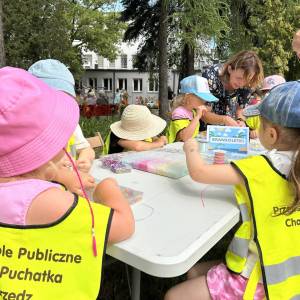 Dzieci z Przedszkola Chatka Puchatka ubrane w kamizelki odblaskowe pracują przy stanowisku z bransoletkami.
