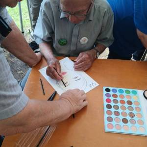 Dwóch seniorów z Dziennego Domu Senior Wigor ozdabia kobiecą twarz na kartce papieru nakładając na nią pędzelkiem kolory. Obok widać paletę z kolorami.