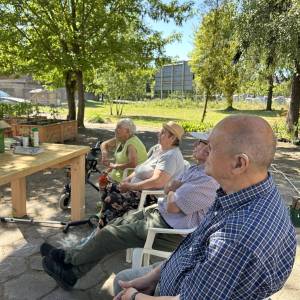 Dwie seniorki i dwóch seniorów siedzący na krzesłach w cieniu słuchają prelegenta, specjalistę od ogrodnictwa.