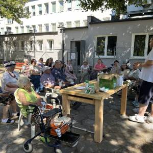 Seniorzy i pracownicy Centrum Usług Społecznych siedzą na krzesłach w cieniu i słuchają opowieści prelegenta. W tle widać budynek Centrum.