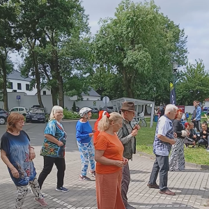 Grupa przebranych seniorów wykonuje układ taneczny na placyku przed wejściem do Ośrodka Kultury. W tle widać drzewa, mur i parking z samochodami.