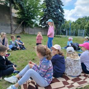 Warsztaty kulinarne Nie marnujemy, wszystko ugotujemy. Zbliżenie na dzieci siedzące w kręgu na trawie. W tle drzewa, mur i brama wjazdowa.