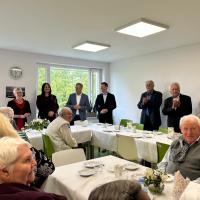 Za uroczystymi stołami siedzą uśmiechnięci seniorzy i seniorki. W tle przy dwóch długich stołach stoją: dyrektor Centrum Usług Społecznych, pierwsza wicewojewoda, burmistrz i jego pierwszy zastępca, przewodnicząca rady miejskiej w Swarzędzu i radni.