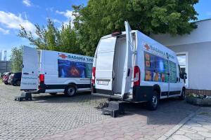Na zdjęciu widać dwa duże busy diagnostyczne stojące na parkingu przy Centrum Usług Społecznych w Swarzędzu. W tle widać drzewa, błękit nieba i chmury.