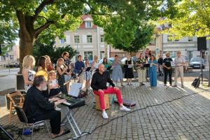 Plac Niezłomnych. Swarzędzka Orkiestra Flażoletowa daje koncert plenerowy na Placu Niezłomnych pod dużym dębem. W tle widać drzewa, kamienice i samochody.