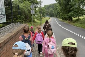 Widok zza uczestników półkolonii na szosę biegnącą przez łąki i las. Grupa prowadzona przez wychowawczynię idzie po chodniku wzdłuż szosy.