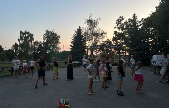 Potańcówka w Gortatowie. Zdjęcie wykonane po zachodzie słońca. W półmroku na placu przy świetlicy wiejskiej kilkanaście osób w różnym wieku tańczy układ. W tle widać drzewa, błękit nieba i łunę po zachodzie słońca.