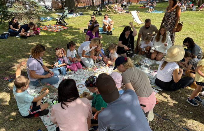 Śniadanie na trawie w parku przed Ośrodkiem Kultury w Swarzędzu. Dookoła koca rozłożonego na trawie klęczą dzieci i ich rodzice i ozdabiają drewniane serca wykorzystując różne elementy dekoracyjne. W tle widać kilkanaścioro innych uczestników pikniku.