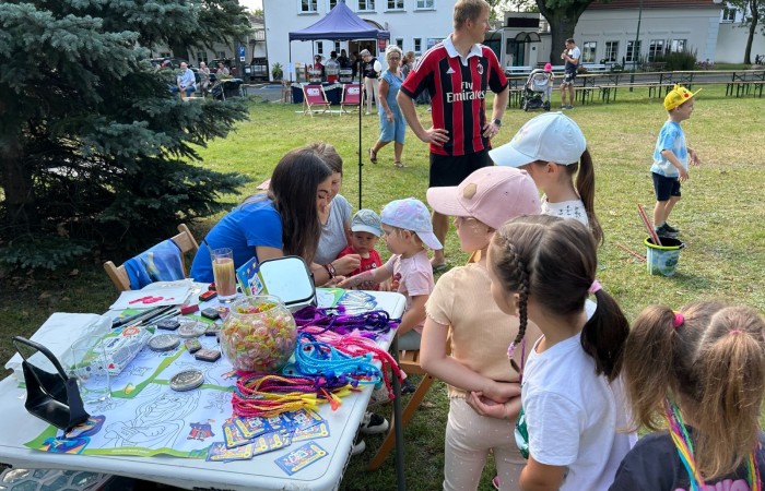 Śniadanie na trawie. Kolejka dzieci czeka do stanowiska, w którym instruktorka z Maxi Fun wykonuje tatuaże i zaplata kolorowe warkoczyki. W tle widać innych piknikowiczów i siedzibę Ośrodka Kultury.