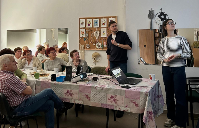 Wnętrze głównej sali w Centrum Aktywności Seniora przy Placu Handlowym w Swarzędzu. Wolontariuszka Europejskiego Korpusu Solidarności, Żuliet Żama, opowiada o swojej rodzinie. Po lewej stronie zdjęcia widać seniorki i seniorów siedzących przy stole przybranym ceratą we wzory florystyczne, patrzących w stronę ekranu. Na stole, przed każdym z seniorów stoją kubki z kawą i herbatą. Po prawej stronie zdjęcia stoi koordynator projektu Dariusz Bugajski i wolontariuszka Żuliet. Oboje patrzą w kierunku, z którego pada światło ekranu.