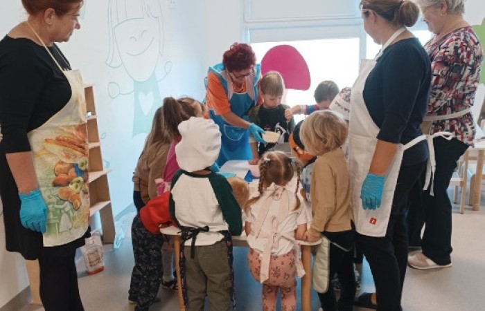 Zdjęcie prezentuje seniorów w towarzystwie dzieci, którzy wspólnie pracują nad kuchennymi wypiekami. Wszystkie osoby są ubrane w kuchenne fartuszki.