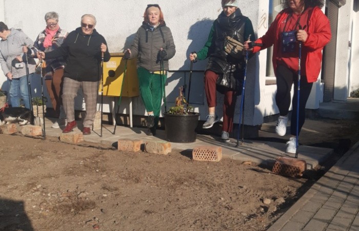 Fotografia przedstawia seniorki w towarzystwie instruktora zajęć, które przygotowują się do wspólnego wymarszu na zajęcia nordic walking. W tle widać budynek Klubu Młodych Duchem Pod Lasem
