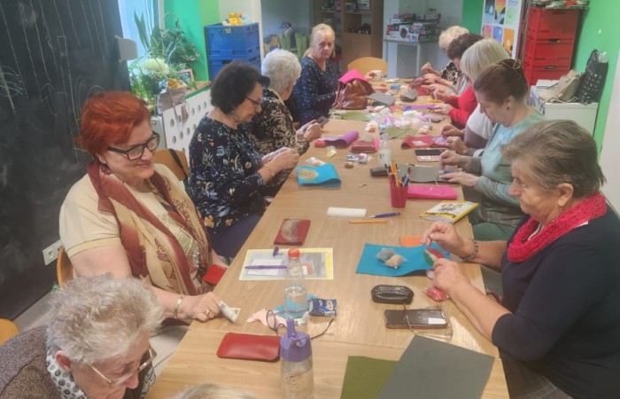 Na zdjęciu widać uśmiechnięte panie, które tworzą kolorowe wyroby artystyczne na zajęciach. W tle mieni się mnóstwo ozdób w przekroju barw tęczy